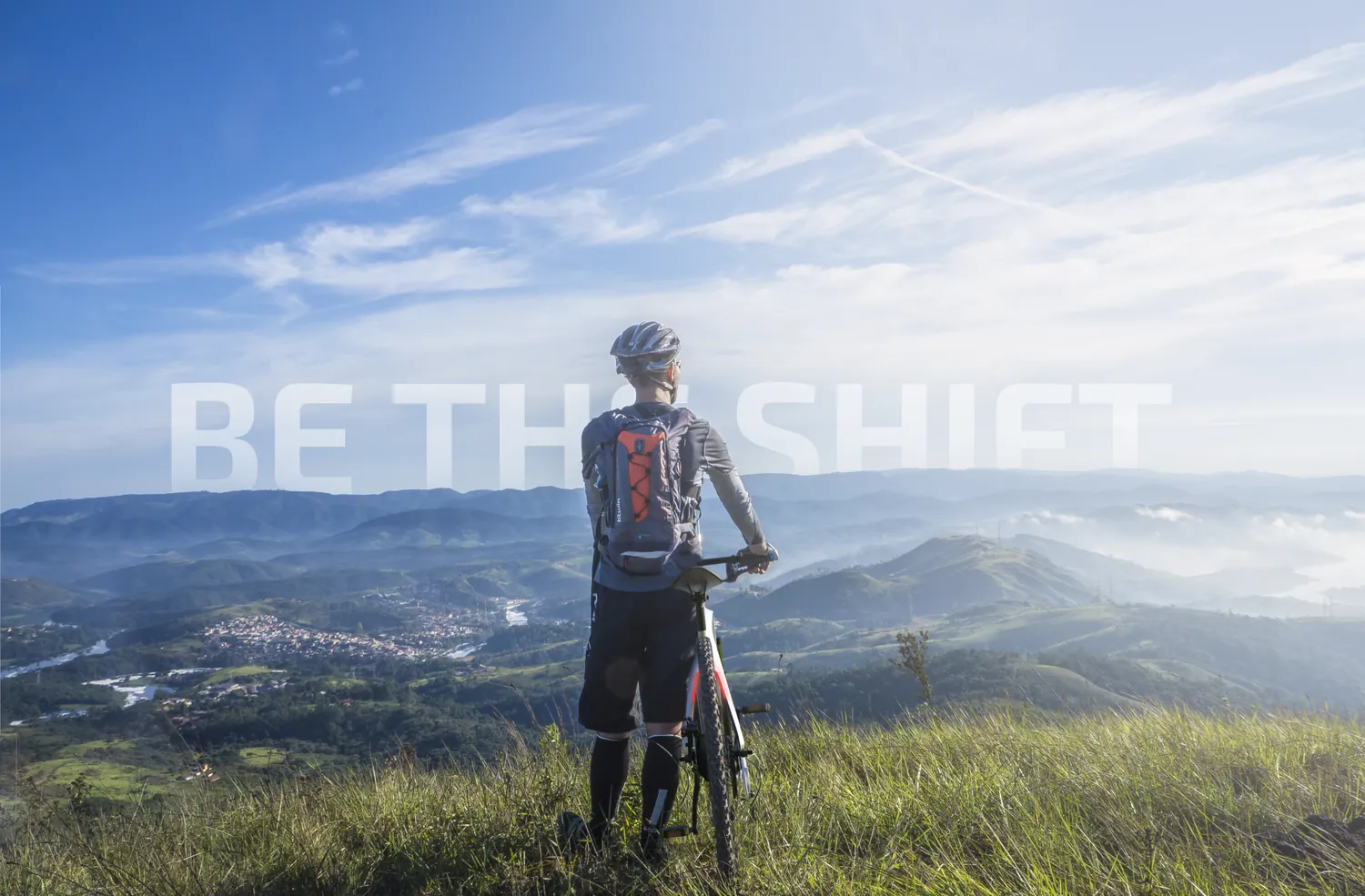 A cyclist standing with a bicycle on a grassy ridge above a valley, with the words Be the shift across the sky.